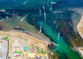 Spot de kitesurf et de l'hôtel Urla Surf House en Turquie avec des kiteurs, des logements, la plage, une eau bleu claire et une vue de drône