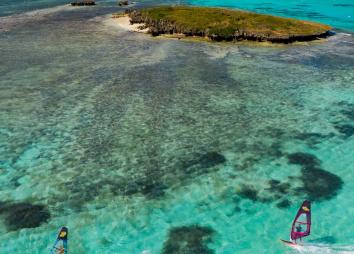 Deux windsurfeurs en action sur l'eau truquoise et transparente du lagon de Madagascar avec une petite île au loin avec du sable blanc, des coraux et de la végétation 