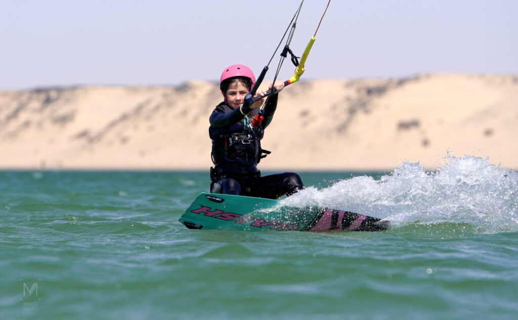 Une jeune fille navigue en kitesurf sur l'eau devant une grande dune de sable.