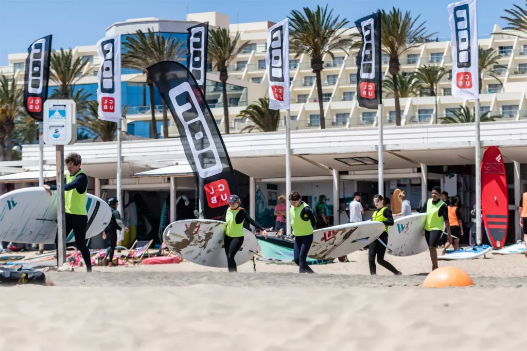 Plusieurs pratiquants en gilet jaune transportent des planches de windsurf devant le centre ION CLUB et des drapeaux sur la plage de Fuerteventura.