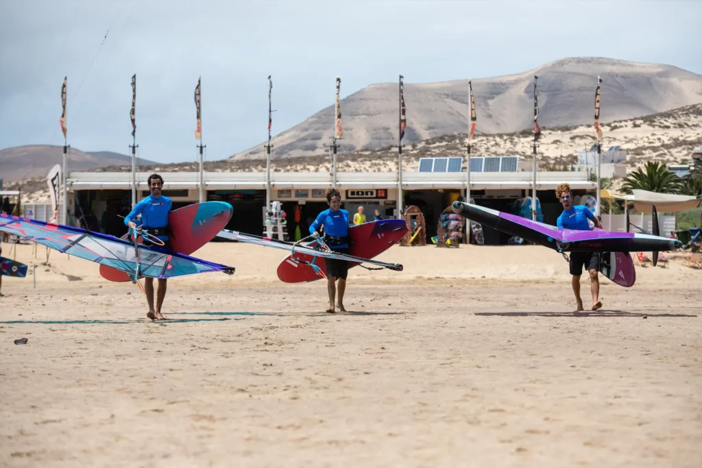 Trois moniteurs de sports de glisse marchant sur la plage de Fuerteventura avec du matériel de windsurf et de wingfoil.