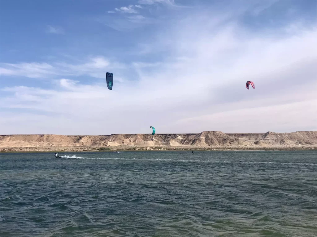 Vue aérienne de trois voiles de kitesurf colorées (rose, bleue et jaune) survolant une immense plage de sable blanc et un lagon à marée basse à Zanzibar.