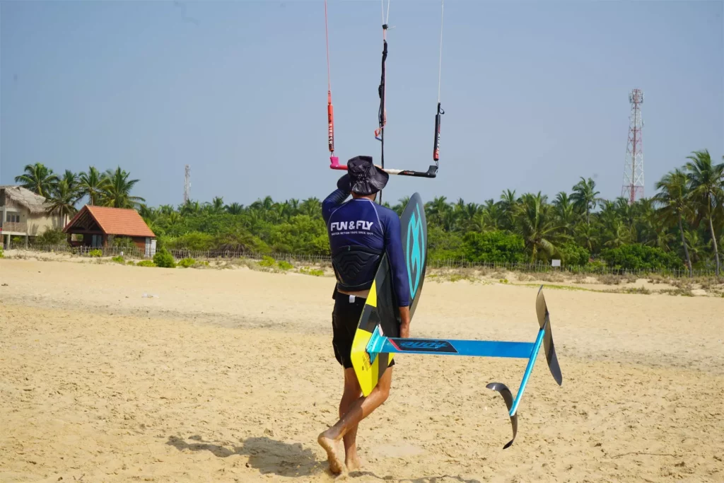 Un homme portant un t-shirt Fun & Fly marche de dos sur une plage de sable en tenant une barre de kitesurf et une planche de kite foil F-One à Kalpitiya au Sri Lanka.
