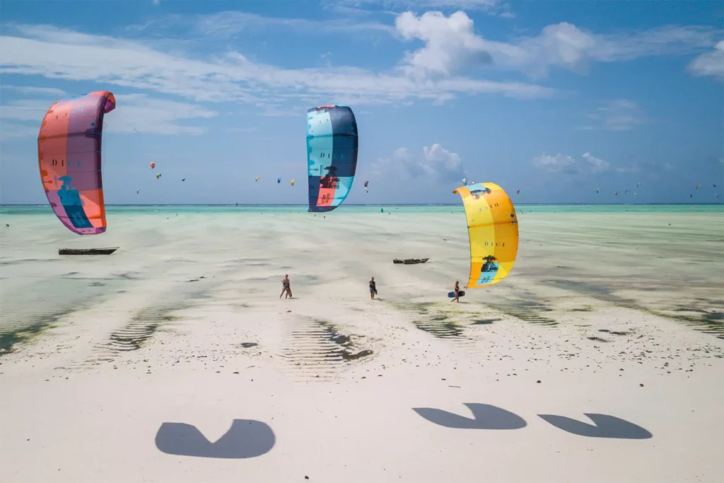 Vue aérienne de trois voiles de kitesurf colorées (rose, bleue et jaune) survolant une immense plage de sable blanc et un lagon à marée basse à Zanzibar.