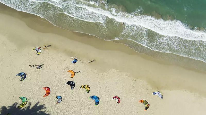 Vue aérienne de haut de voiles de kitesurf multicolores disposées sur le sable fin d'une plage en bord de mer.