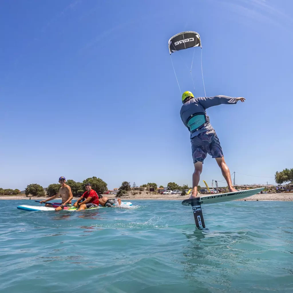 Un kitesurfeur s'élève au-dessus de l'eau en kite foil avec une aile Core sous un ciel bleu à Rhodes, tandis que des personnes l'observent depuis un stand up paddle.
