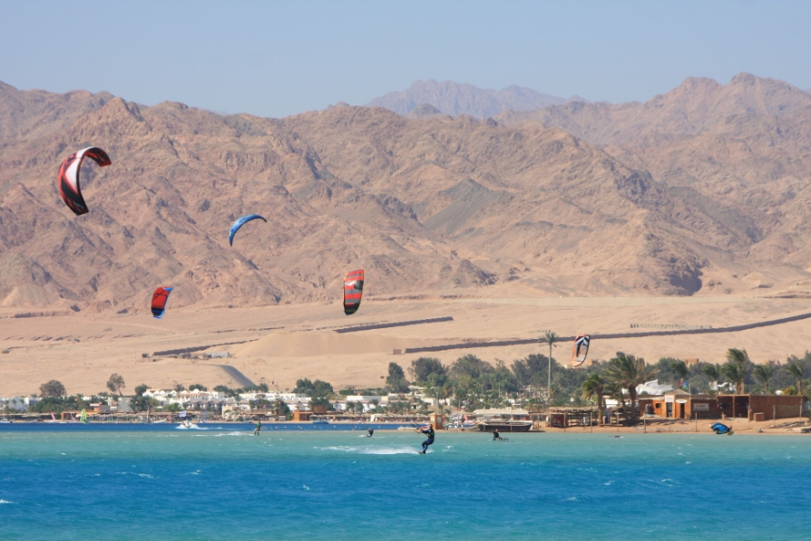 Plusieurs kitesurfeurs naviguent sur une mer bleue face à des montagnes désertiques à Dahab en Égypte.