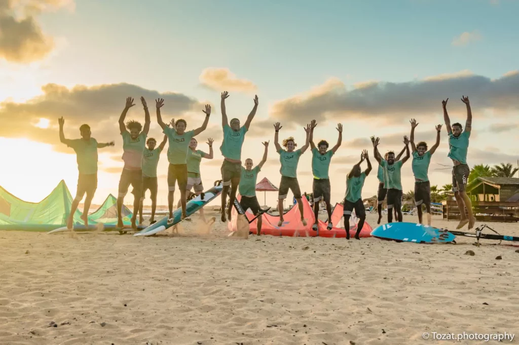 Plusieurs pratiquants en gilet jaune transportent des planches de windsurf devant le centre ION CLUB et des drapeaux sur la plage de Fuerteventura.