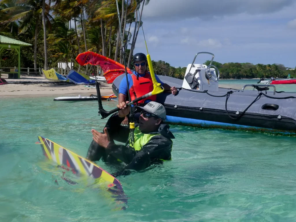 Un moniteur en gilet rouge sur un bateau semi-rigide encadre un pratiquant de kitesurf dans une eau transparente à Sainte-Anne en Guadeloupe.