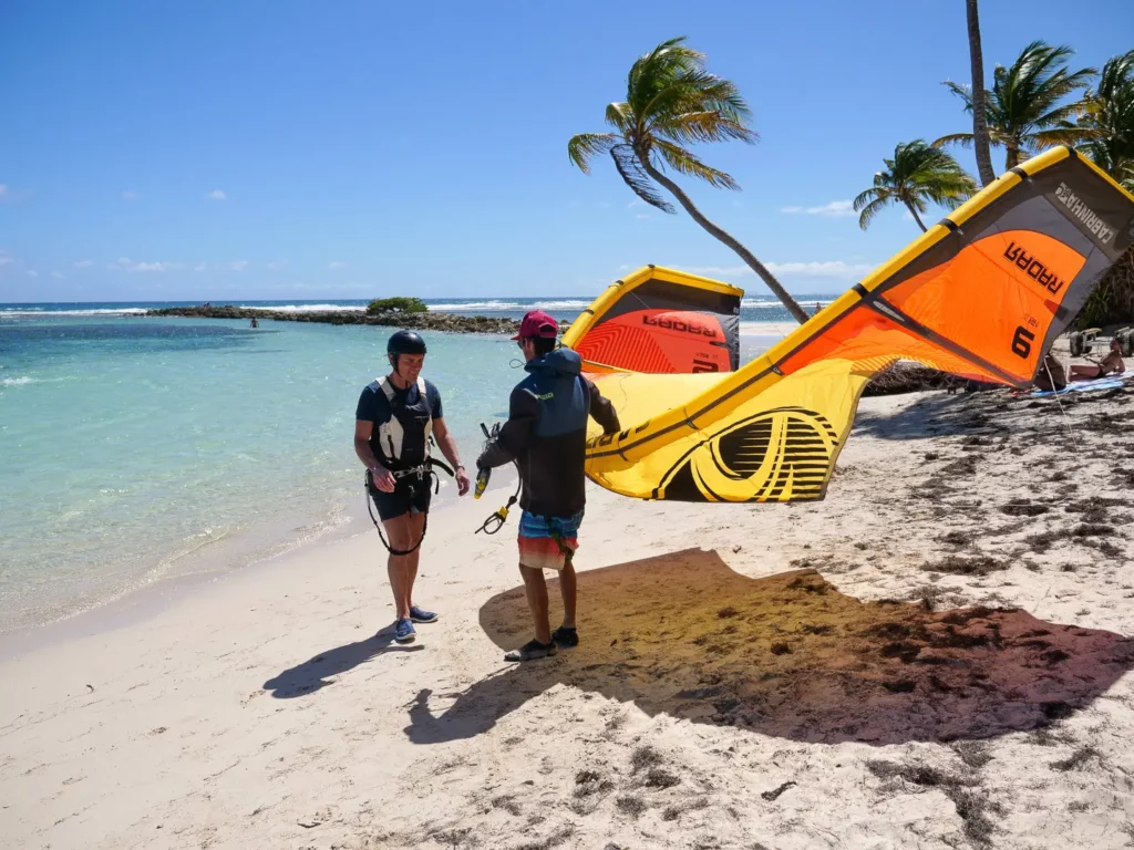 Un moniteur prépare une aile de kitesurf jaune et orange Cabrinha Radar sur une plage de sable blanc bordée de palmiers et d'un lagon.