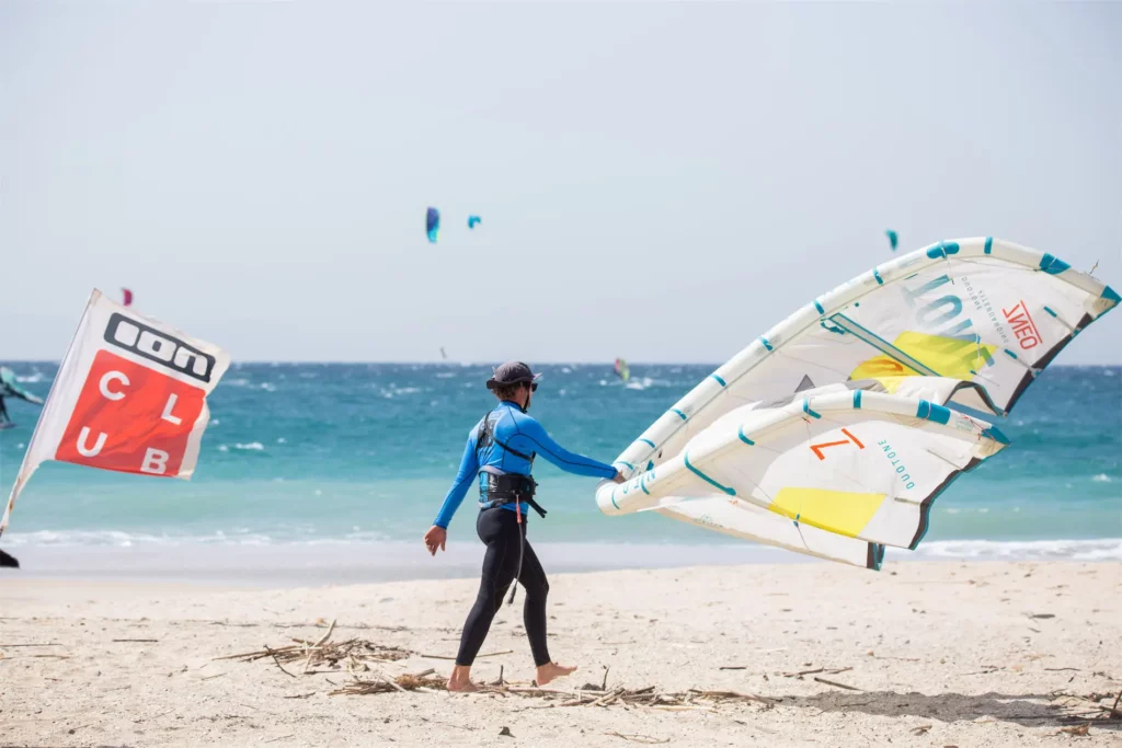 Un kitesurfeur en combinaison bleue marche sur le sable en portant une aile de kitesurf Duotone Neo blanche et jaune près d'un drapeau ION CLUB.