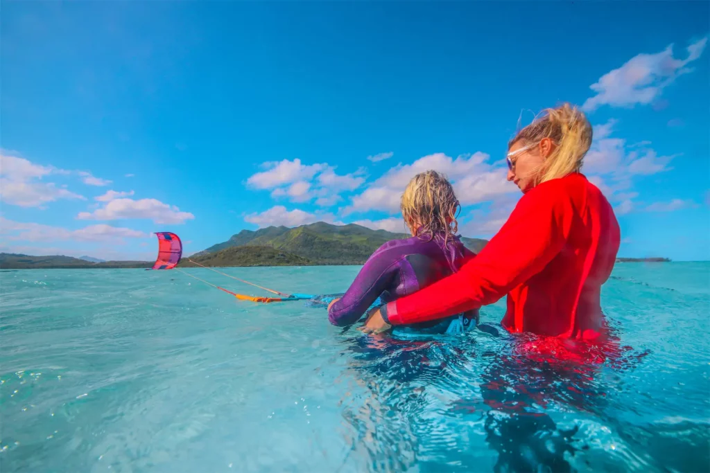 Un moniteur en t-shirt orange conseille un élève en gilet et casque tenant sa barre de kitesurf dans l'eau turquoise en Grèce.