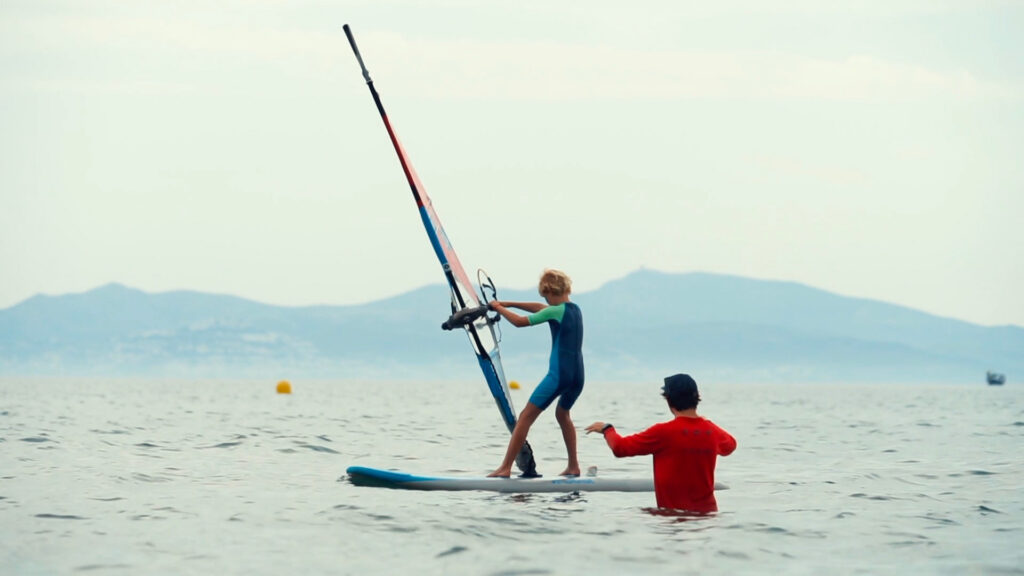 Un enfant apprend la planche à voile (windsurf) avec un moniteur dans l'eau.