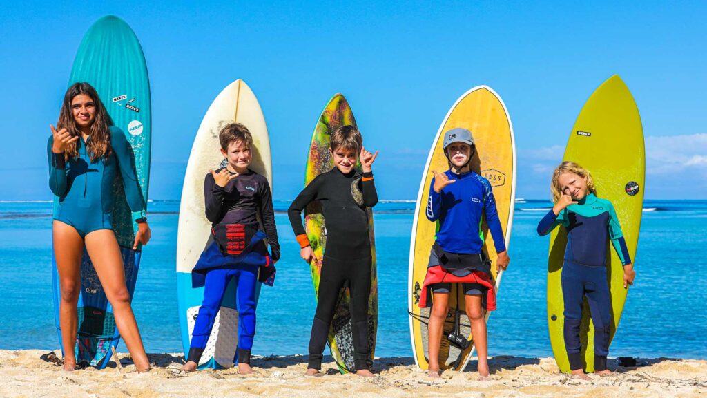 Cinq enfants en combinaison de surf posant sur la plage avec leurs planches devant une mer turquoise.