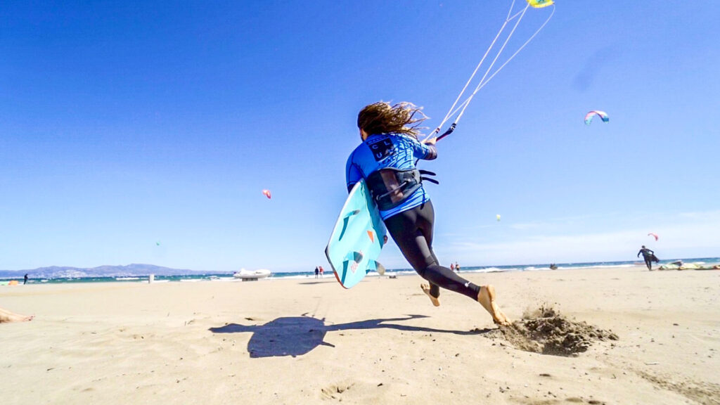 Un kitesurfeur en combinaison bleue court sur le sable d'une grande plage avec sa planche pour se lancer à l'eau à Rosas en Espagne.